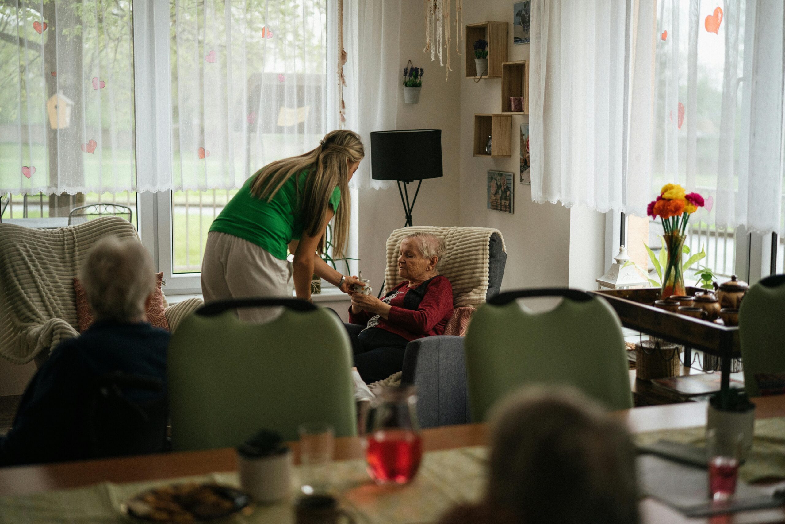 Caregiver assisting elderly women with tea in a cozy nursing home setting.